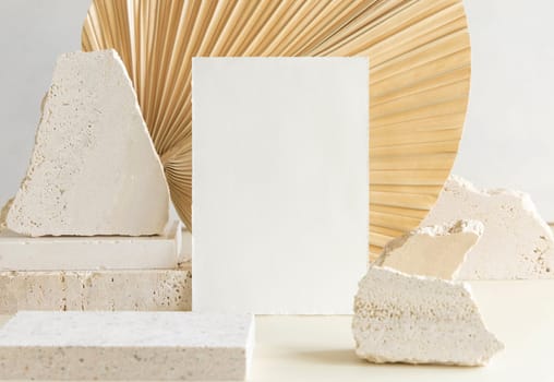Blank card near beige travertine stones against dry beige palm leaf close up, mockup. Boho scene with vertical paper card. Bohemian or close to nature greeting or wedding Invitation. Minimal composition.