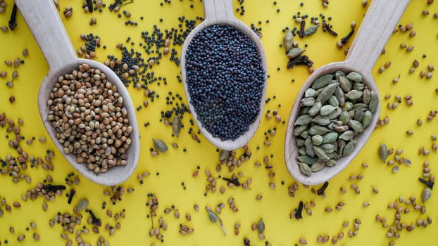 Black mustard seeds, coriander and cardamom in wooden spoon on yellow background from above