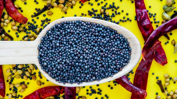Wooden spoon with black mustard seeds on yelow background with red dry peppers