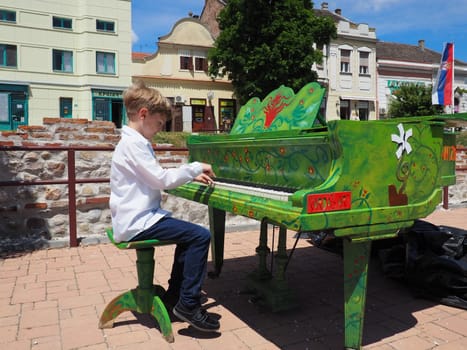 Sremska Mitrovica, Serbia, 05 June 2021. A boy in a white shirt plays the piano in the street. The child is 9 years old Caucasian. Piano painted green. Performance of a piece of music. Street concert.