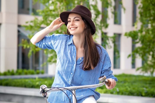 Pretty smiling girl in hat and blue dress riding a bicycle at street lifting legs