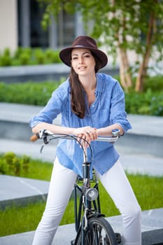 Pretty smiling girl in hat and blue dress riding a bicycle at street lifting legs