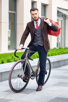 Handsome businessman in a jacket with red bag sitting on  his bicycle on city streets. The concept of the modern lifestyle of young men