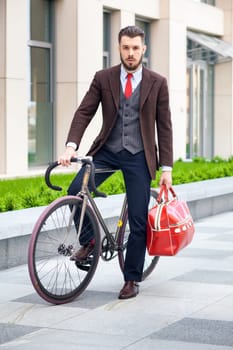 Handsome businessman in a jacket with red bag sitting on  his bicycle on city streets. The concept of the modern lifestyle of young men