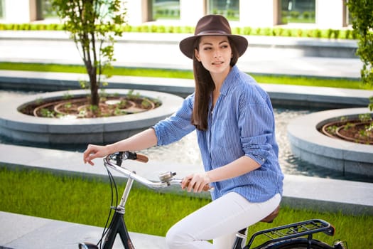Pretty smiling girl in hat and blue dress riding a bicycle at street