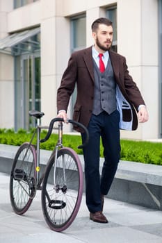 Handsome businessman in a jacket and red tie and his bicycle on city streets. The concept of the modern lifestyle of young men