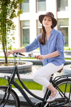Pretty smiling girl in hat and blue dress riding a bicycle at street