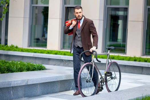 Handsome businessman in a jacket and red tie and his bicycle on city streets. The concept of the modern lifestyle of young men