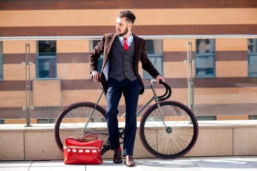 Handsome businessman in a jacket and red tie and his bicycle on city streets. red bag lies next. The concept of the modern lifestyle of young men