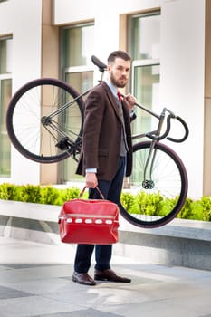 Handsome businessman with a red bag carrying his bicycle on city streets. The concept of the modern lifestyle of young men