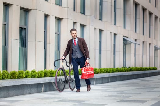 Handsome businessman in a jacket and red bag and his bicycle on city streets. The concept of the modern lifestyle of young men