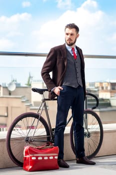 Handsome businessman in a jacket and red tie and his bicycle on city streets. red bag lies next. The concept of the modern lifestyle of young men