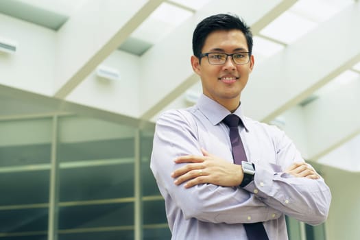 Portrait of happy and confident young asian businessman. The man stands in a street against office buildings and crosses arms looking at camera