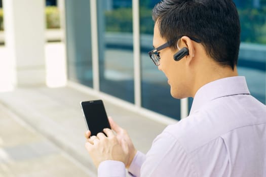 Young chinese businessman typing phone number on smartphone and talking with bluetooth headset device