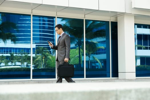 Young chinese businessman commuting to office with computer bag and reading message on mobile phone. The man types while walks