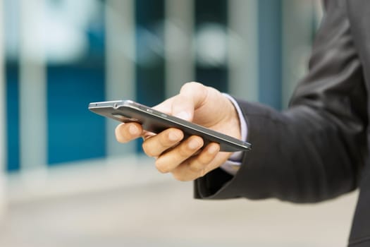 Closeup of businessman typing note on mobile phone. The man writes on his smartphone using one hand with office building in background.