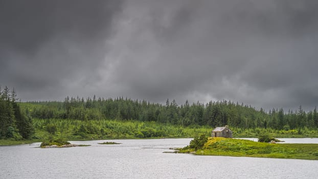 Small fishing house on the edge of Ballynahinch Lake. Pine forest with dark storm sky in background. Connemara, County Galway