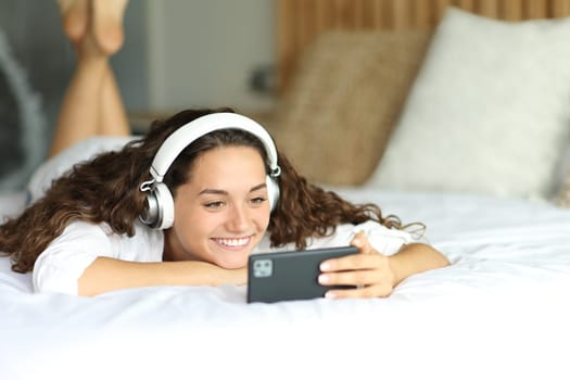 Happy woman wearing headphones watching videos on smart phone lying on a bed in a bedroom