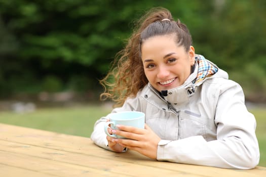Woman in a picnic table drinking coffee in the mountain looking at camera