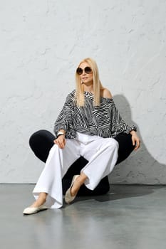 Stylish blonde woman in striped blouse and wide white trousers resting in modern chair under harsh light