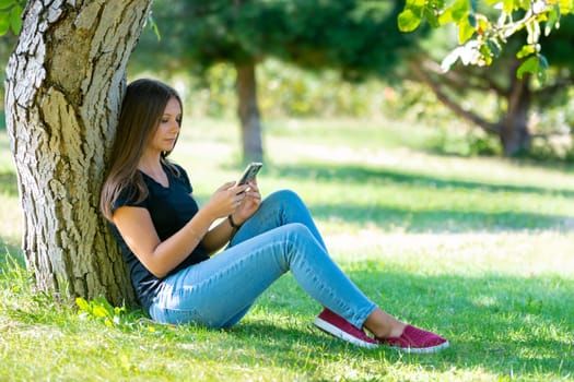 A girl sits under a tree in a sunny park and looks at the smartphone screen