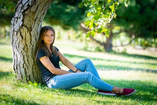 A girl sits under a tree in a sunny park and looks into the frame