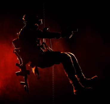 Silhouette of police officer in tactical gear descending from a height, rope exercises with weapons. Tactical rappelling, anti-terror or counter terrorism operation in darkness in rappelling harness