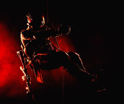 Silhouette of police officer in tactical gear descending from a height, rope exercises with weapons. Tactical rappelling, anti-terror or counter terrorism operation in darkness in rappelling harness