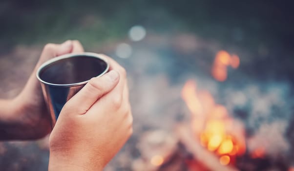 Human's hands holding the metal cup with hot drink above the fire. Concept of the camping and recreation.