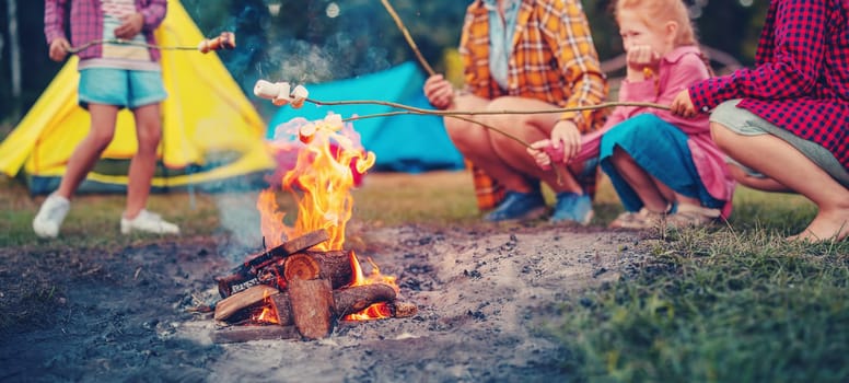 Group of the people sitting near the campfire and cooking marshmallows. Concept of the scouts camping and family vacation.