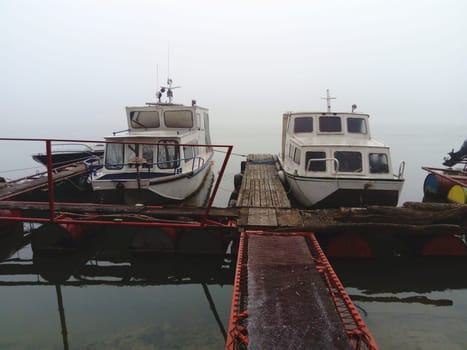 Belgrade, Serbia, January 14, 2020 Two white boats at a wooden pier, left for the winter. Foggy weather in southern Europe. January cloudy day. Reflection of water transport in a river river.