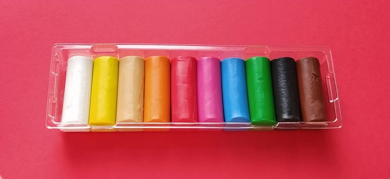 Multi-colored new plasticine in plastic packaging on a red background. Plasticine plates of red, pink, green, white, yellow, brown, blue, black. Set for children's creativity.