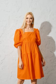 A beautiful woman with blond hair stands in an orange sundress, smiling brightly and looking into the camera