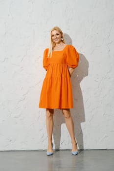 A smiling woman with her hands behind back in a bright orange spring dress stands indoors, posing for the camera