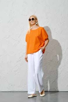 An adult woman walking in a stylish orange and white ensemble, holding a pair of sunglasses. A fashionable portrait perfect for the spring season.