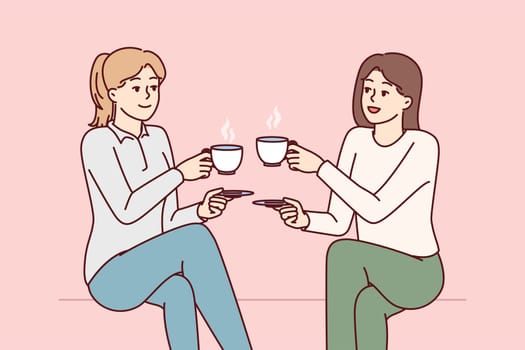 Two beauty girl friends drink hot coffee holding cups and saucers enjoy joint coffeebreak. Ladies dressed in casual style gossip during lunch break with hot non-drinks in hands
