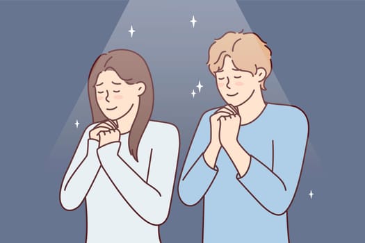Positive man and woman praying asking god for happy marriage. Young couple stand with eyes closed and clasping palms in front of chest together performs religious ritual