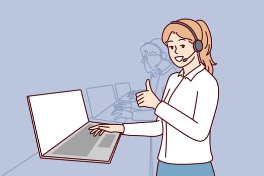 Woman in headphones for call center stands near laptop and looks at camera showing thumbs up. Girl employee of psychological support service or software user consultation smiling