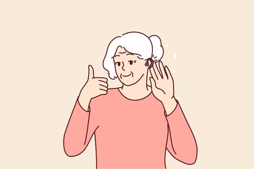 Elderly woman uses hearing aid and shows thumbs up confirming good sound volume thanks to new device. Gray-haired old lady with hearing aid enjoys opportunity to hear others and communicate freely