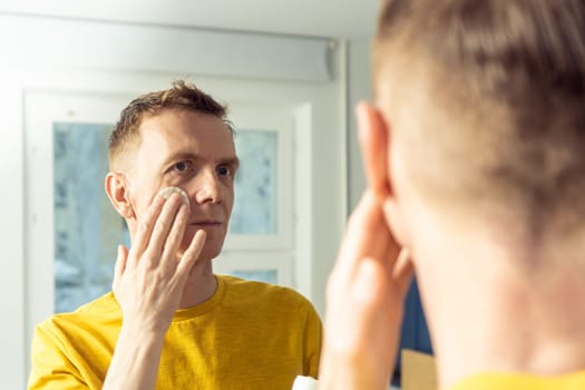 Middle aged man apply face tonic with cotton pad, then massage with hands. Male portrait in mirror. Skincare morning routine, careful beauty treatment, home lifestyle. Face cleansing, washing.