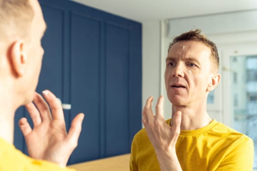 Middle aged man apply moisturizing cream with finger on clear facial areas. Male portrait in room mirror. Skincare morning routine, careful beauty treatment, home lifestyle. Facial care concept.