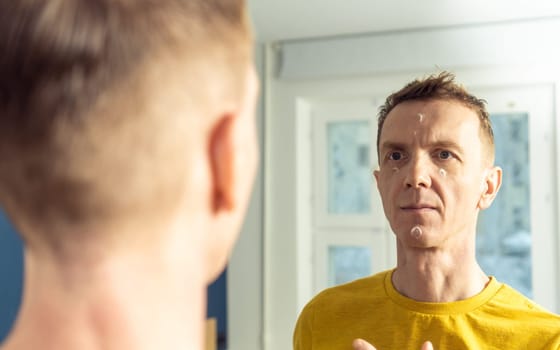 Middle aged man apply moisturizing cream on clear facial areas. Male portrait in room mirror. Skincare morning routine, careful beauty treatment, home lifestyle. Facial care concept.