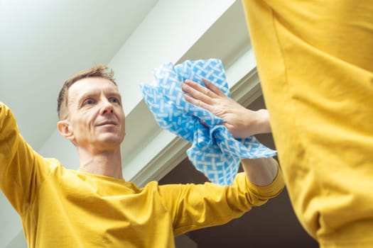 Mature man wiping room mirror with rag, from below view. Happy man rub from dust and polish mirror with household cloth. Domestic life, tidy housekeeper. Housework, home cleaning concept.