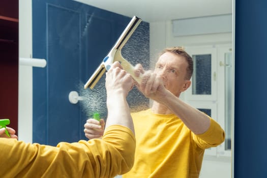 Middle aged man spray cleanser on mirror of sliding wardrobe door and wipe with plastic brush. Rub from dust and polish mirror with household tool. Housework, wet home cleaning concept.