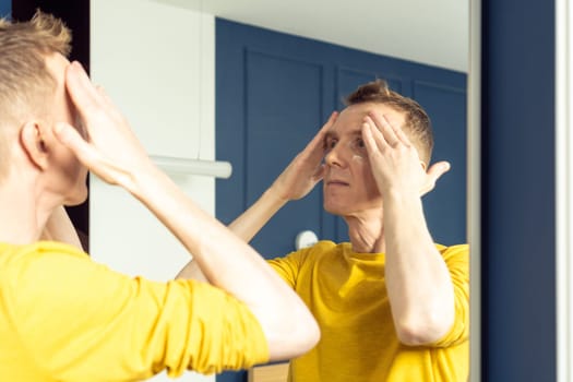 Middle aged man apply cream on clear face, then massage with hands. Male portrait in room mirror. Skincare morning routine, careful beauty treatment, home lifestyle. Facial care concept.