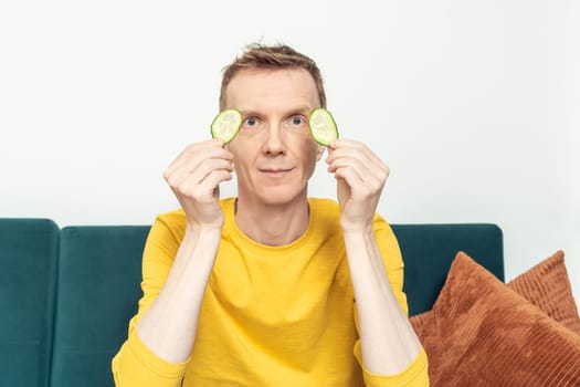Happy adult man sitting on sofa hold in hands eye patches from fresh cucumber slices in living room. Mature person do moisturizing mask for dark circles under eyes. Home lifestyle, beauty treatment.