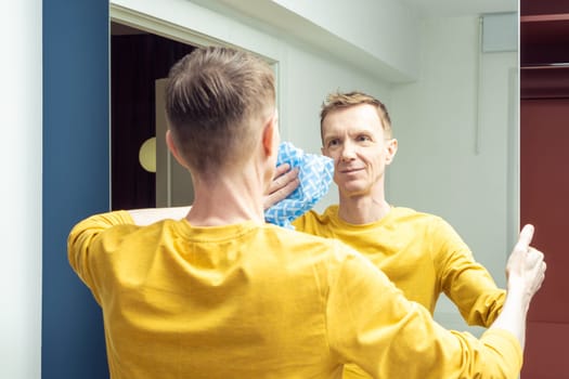 Happy adult man wiping mirror of sliding wardrobe door with dry rag, back view. Rub from dust and polish mirror with household cloth. Domestic life, tidy room. Housework, home cleaning concept.
