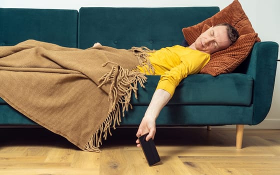 Tired adult man sleep on sofa with smartphone in hanging hand in living room. Overworking person reclining on pillow under blanket, side view. Home relaxation, rest break, leisure, lounge.