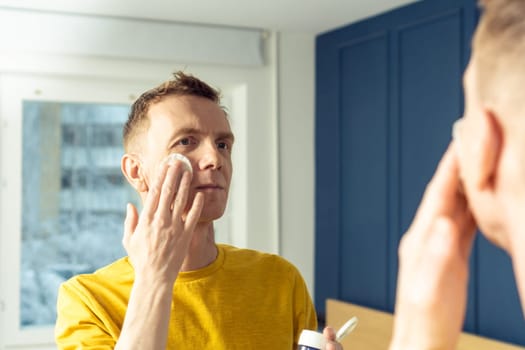 Mature man apply face tonic with cotton pad, then massage with hands. Male portrait in room mirror. Skincare morning routine, careful beauty treatment, home lifestyle. Face cleansing, washing.
