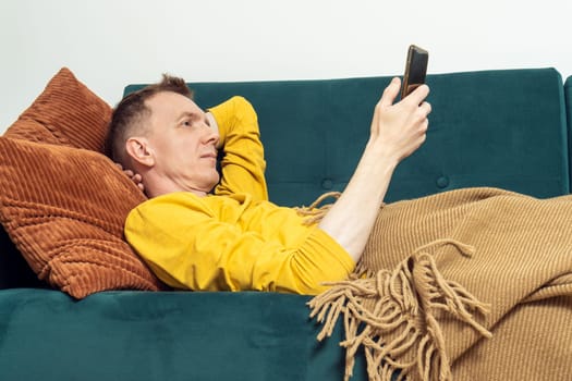 Adult man lie on sofa and watch video on smartphone in living room, side view. Person reclining on pillow under blanket and lounging. Look at screen of phone. Home relaxation, leisure, lazy activity.